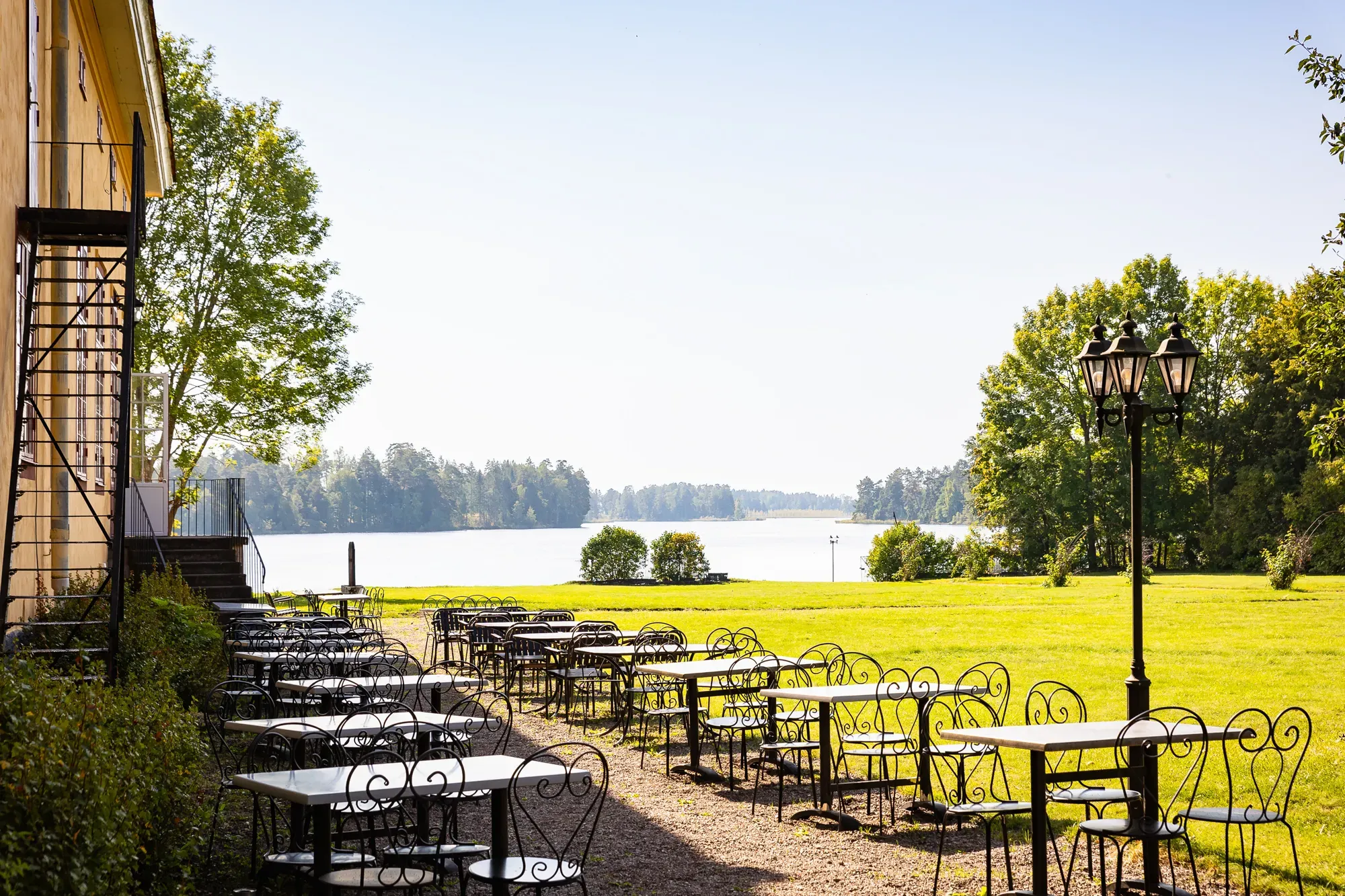 Smidesmöbler på grusgången utmed herrgården, med gräsmattan och Dalälven bakom.